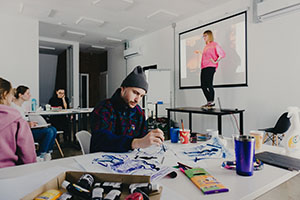 student in foreground painting with teacher/student standing on desk in front of projector screen in background