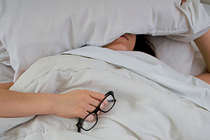 a person is sleeping underneath a pillow. their body is covered by the cover and their arm is exposed. They are holding a pair of black glasses.