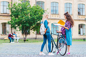 two women conversing in front of a school. One is holding a bike.